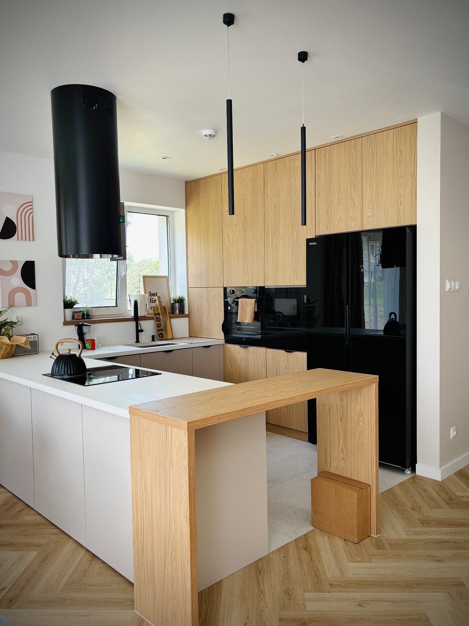Nowoczesna kuchnia z drewnianymi szafkami, czarnym okapem cylindrycznym i wyspą z blatem. Parkiet w jodełkę, minimalistyczne lampy wiszące i czarna lodówka.
