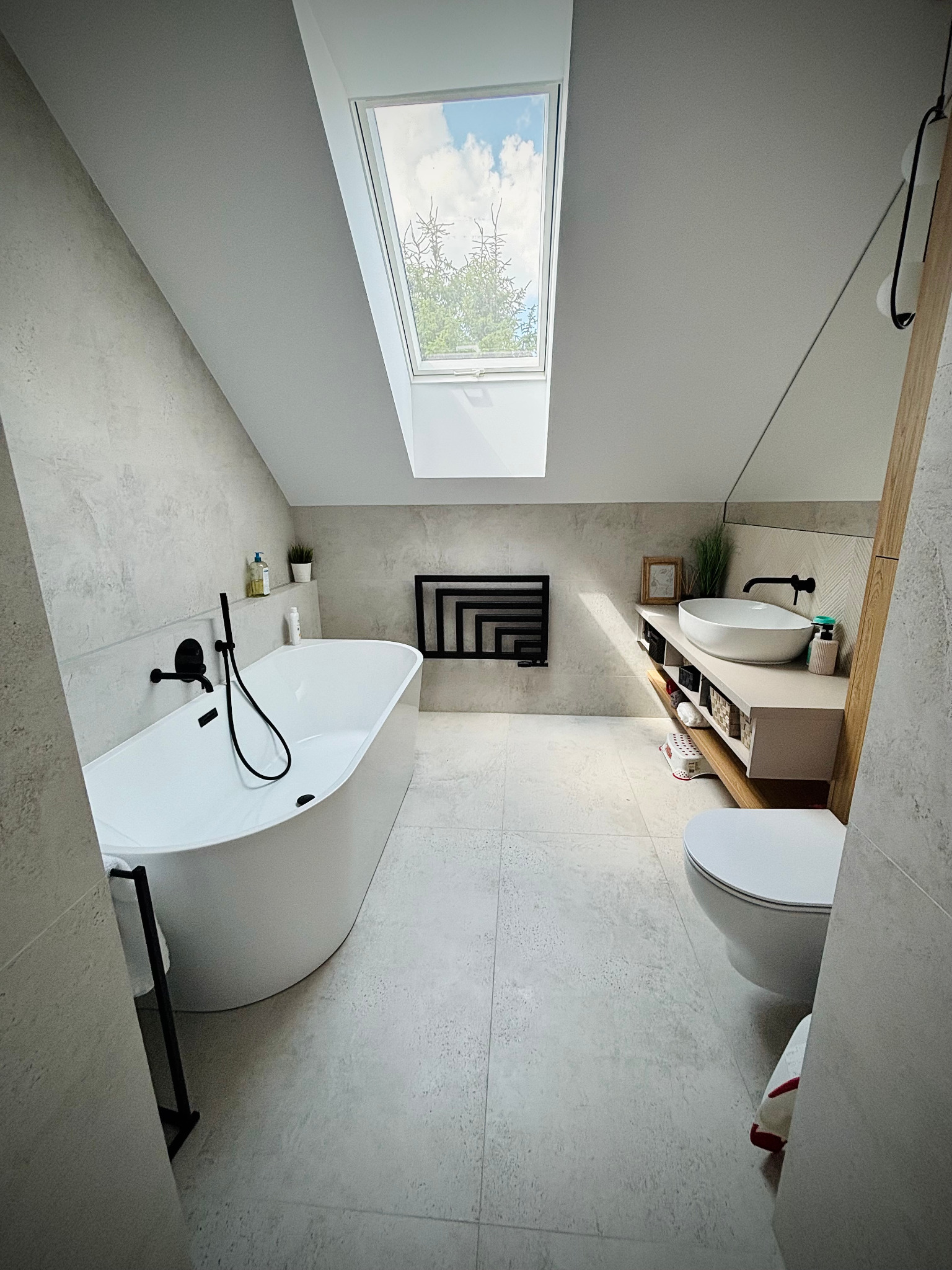 Nowoczesna łazienka na poddaszu z oknem dachowym, białą wanną wolnostojącą i umywalką nablatową. Szaro-biała kolorystyka z czarnymi akcentami.