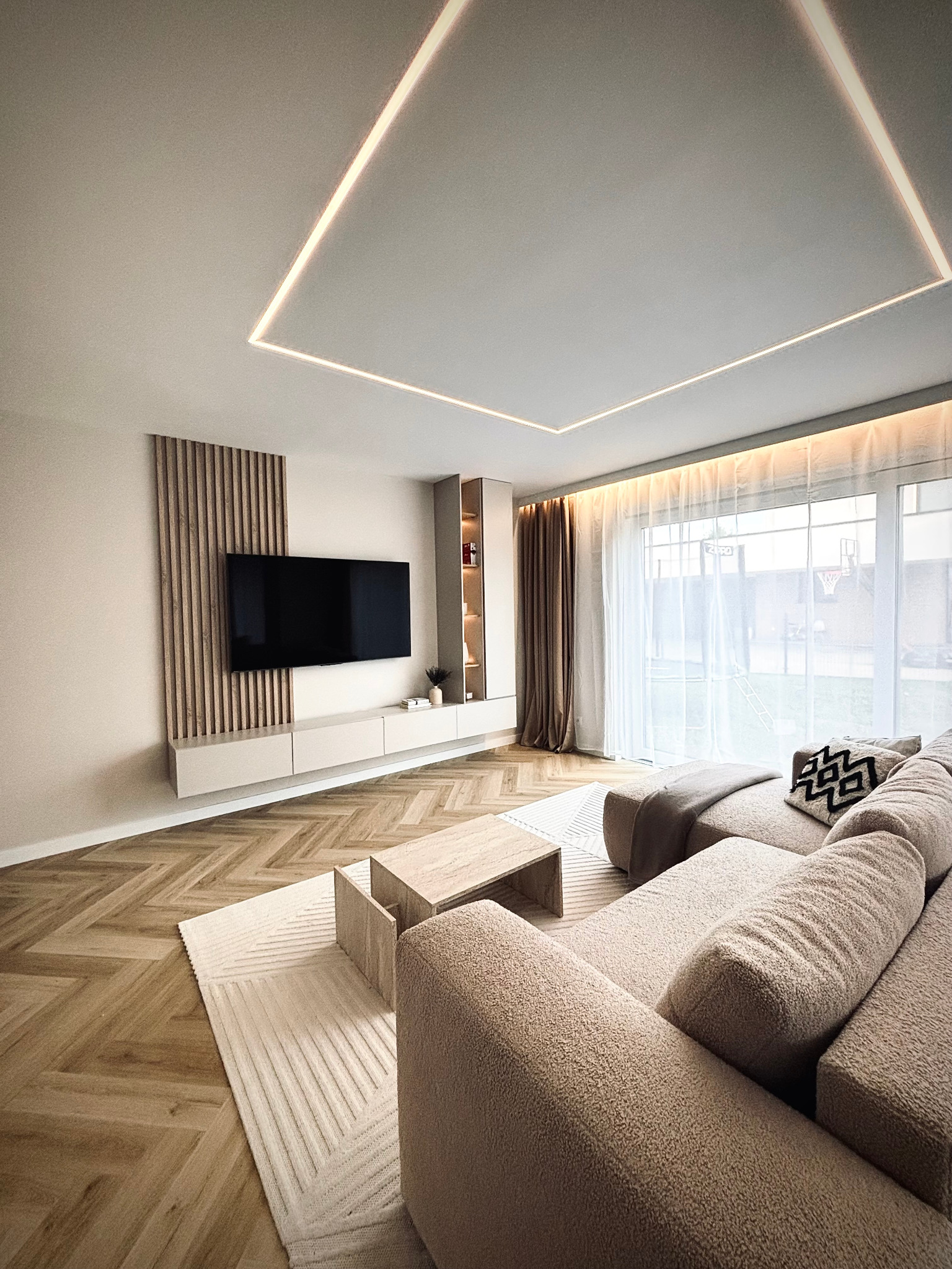 Nowoczesny salon z parkietem w jodełkę, beżową sofą i podświetlanym sufitem. Telewizor na ścianie z drewnianymi lamelami. Minimalistyczny stolik kawowy.