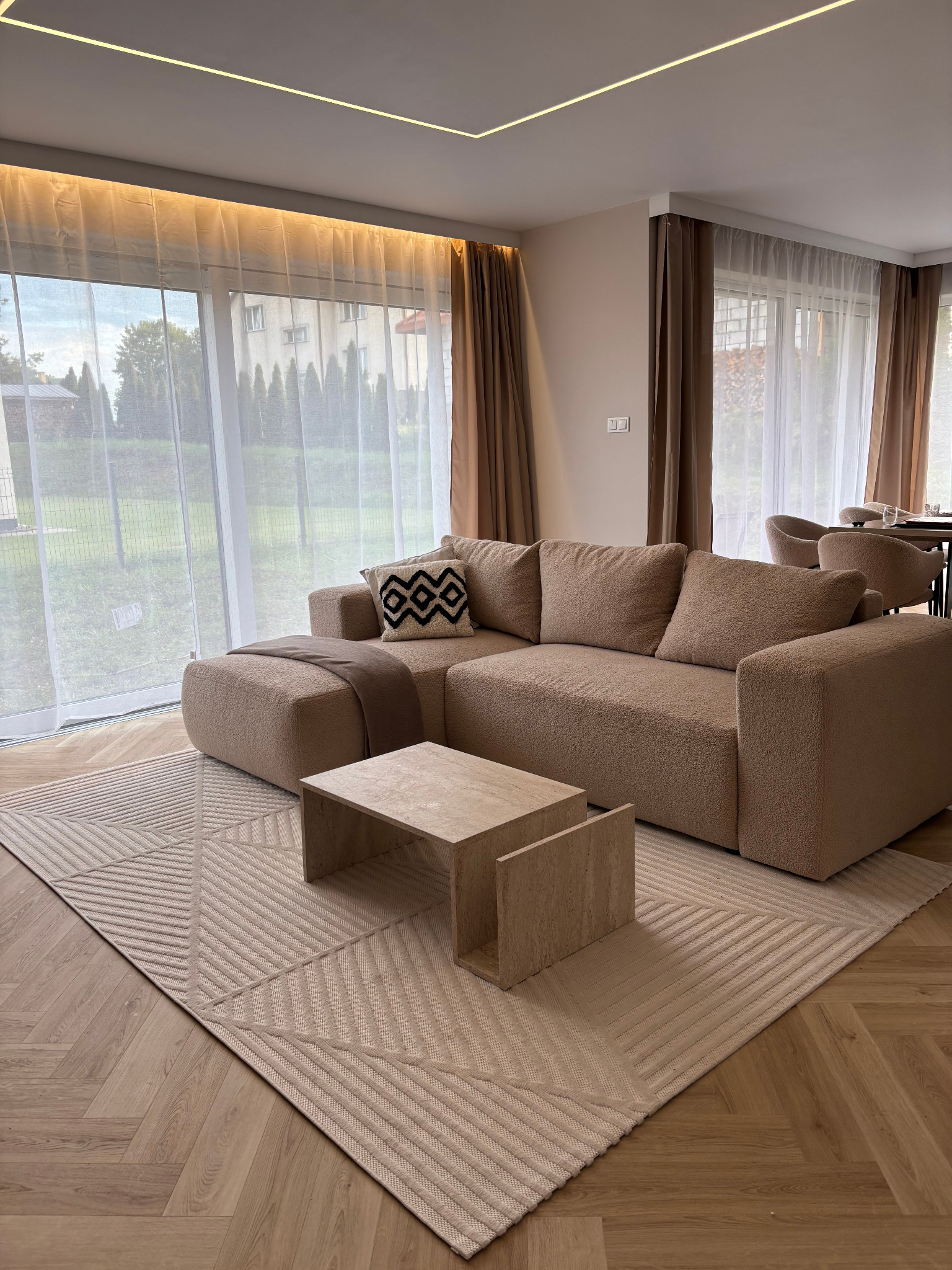 Beżowy narożnik z poduszką we wzory, stolik z trawertynu i dywan w geometryczne wzory na parkiecie ułożonym w jodełkę. W tle okno z jasnymi zasłonami i widok na zieleń.