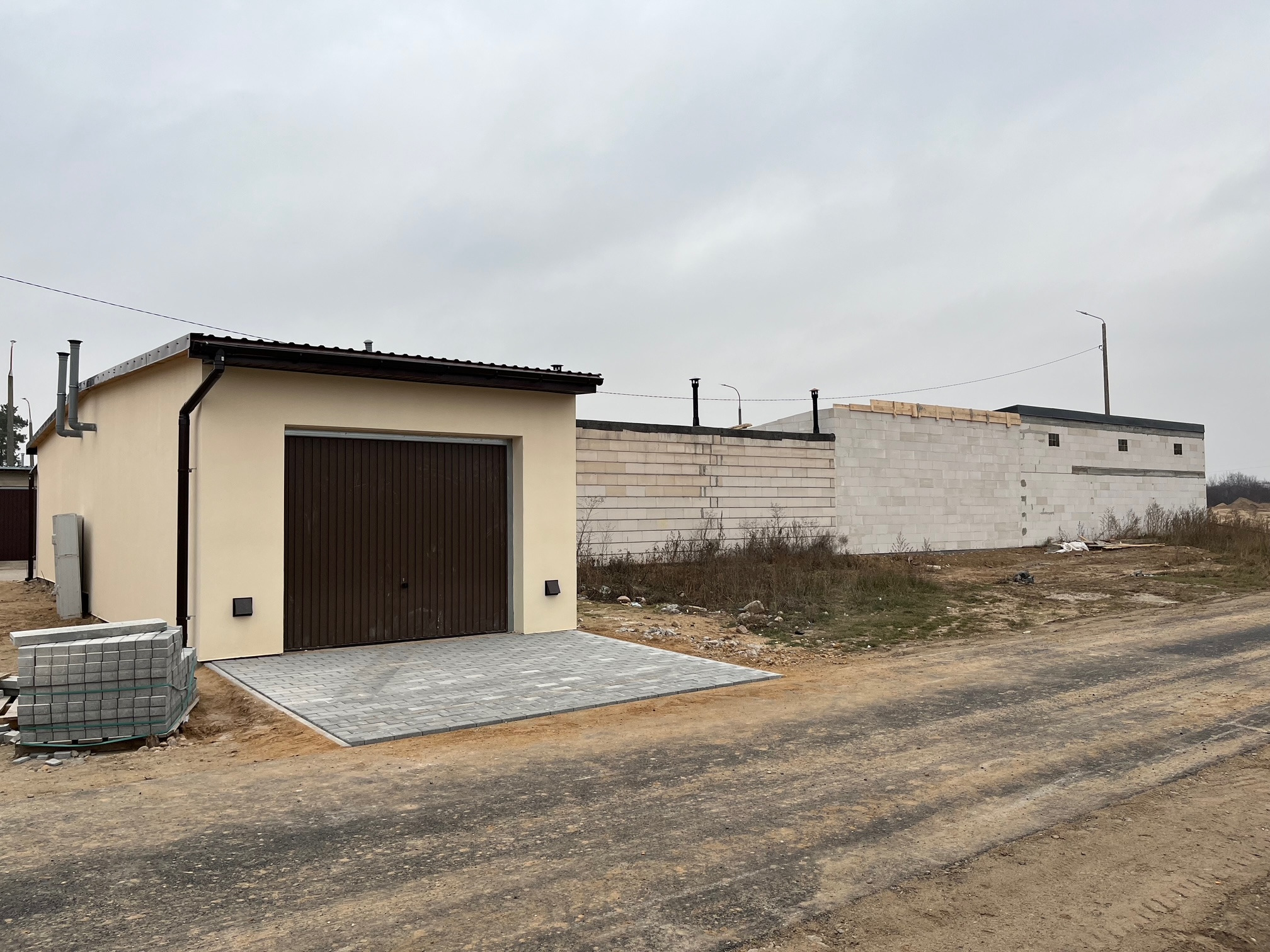 Garaż z brązową bramą i ułożoną szarą kostką brukową. Obok stos kostki. W tle budynek w trakcie budowy z białego bloczka.