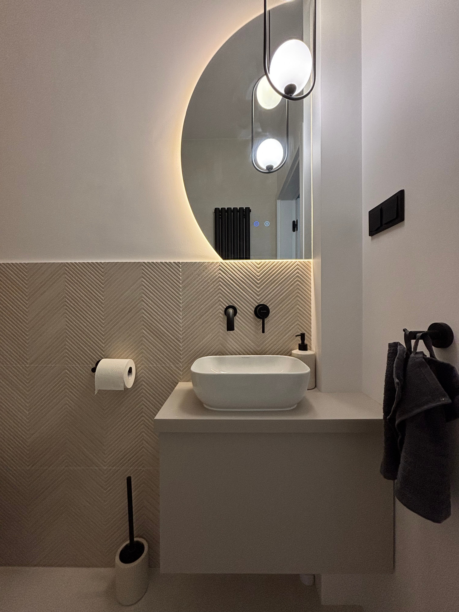 Nowoczesna łazienka z umywalką nablatową, lustrem z podświetleniem LED i beżowymi płytkami ułożonymi we wzór jodełki. Minimalistyczny design, czarne akcenty.