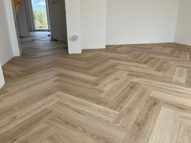 Jasny parkiet ułożony we wzór jodełki francuskiej w nowym, przestronnym wnętrzu. Widok na sąsiednie pomieszczenie przez otwarte drzwi.