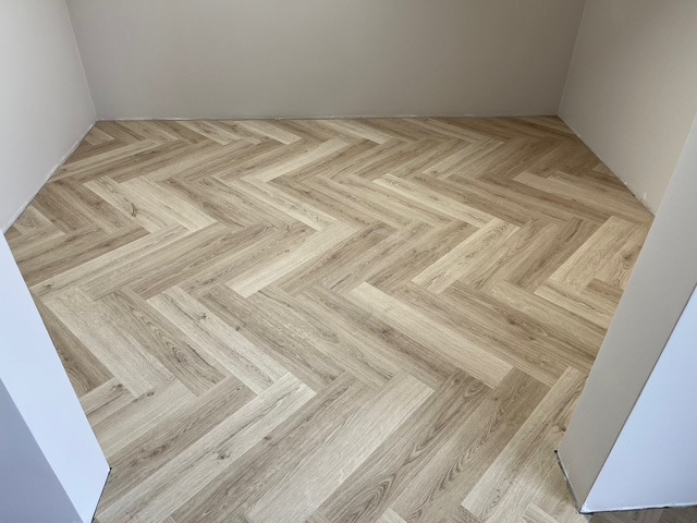 Ułożony parkiet w jodełkę w jasnym odcieniu drewna, widoczne fragmenty ścian w neutralnym kolorze. Całość tworzy estetyczne i schludne wykończenie podłogi.
