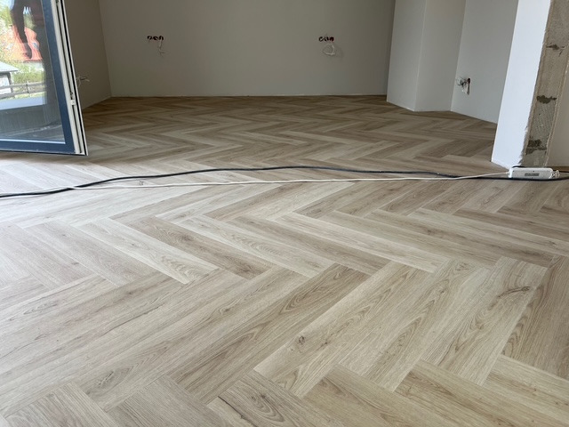 Jasny parkiet ułożony w jodełkę francuską w nowym wnętrzu. Widoczne kable i niedokończone ściany. Duże okno balkonowe po lewej stronie kadru.