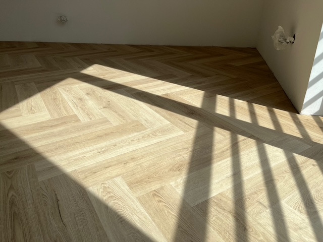 Jasny parkiet ułożony w jodełkę, oświetlony promieniami słońca wpadającymi przez okno. Widoczne białe ściany i wystający kabel elektryczny.