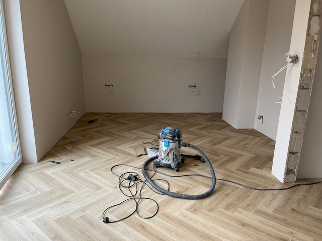 Układanie parkietu w jodełkę w jasnym pokoju na poddaszu. Widoczny odkurzacz przemysłowy i fragment ściany w trakcie wykończenia.