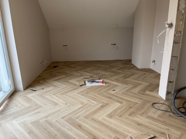 Jasny parkiet ułożony we wzór jodełki w pokoju na poddaszu. Widoczne narzędzia i niedokończone fragmenty podłogi, okno z lewej strony.