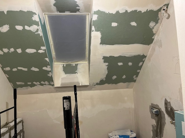 Poddasze w trakcie szpachlowania: sufit ze świetlikiem, częściowo pokryty zieloną masą szpachlową, widoczne narzędzia i worki z materiałami.