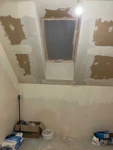 Pokój na poddaszu w trakcie prac przygotowawczych: ściany częściowo pokryte szpachlą, okno dachowe zabezpieczone folią, widoczne worki z zaprawą i narzędzia.