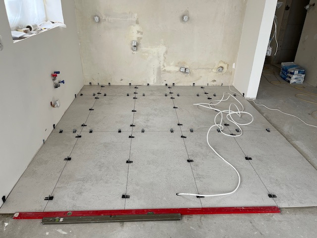 Układanie szarych płytek podłogowych z użyciem krzyżyków dystansowych i poziomicy w jasnym wnętrzu w trakcie remontu. Widoczne instalacje i worki z zaprawą.