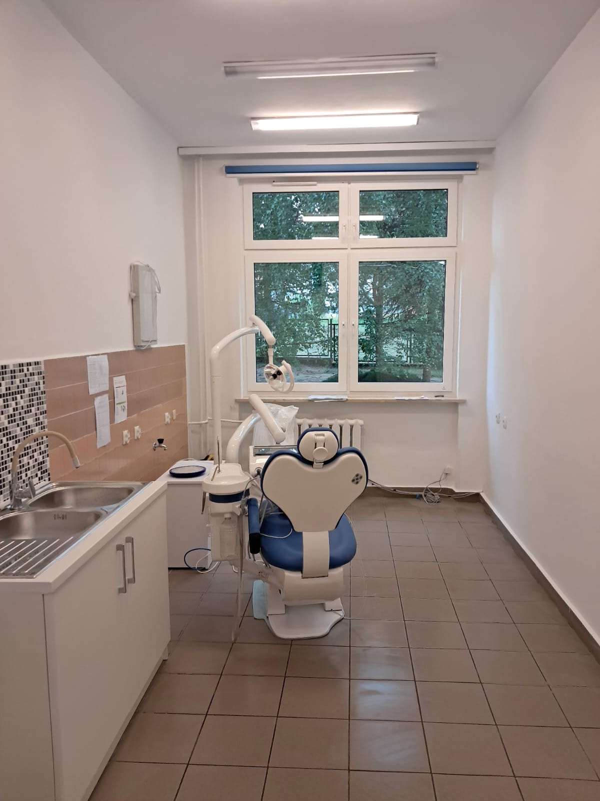 Wnętrze gabinetu dentystycznego z fotelem, umywalką i oknem. Białe ściany, brązowe płytki podłogowe. Widoczne wyposażenie medyczne.