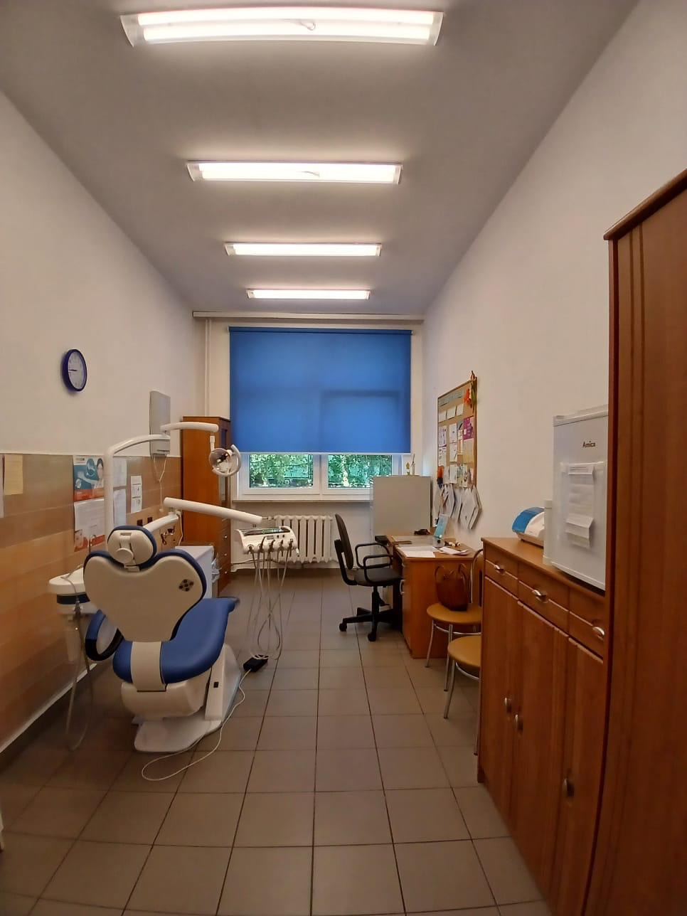 Wnętrze gabinetu dentystycznego z fotelem, biurkiem, szafkami i oknem z niebieską roletą. Widok na stanowisko pracy stomatologa.
