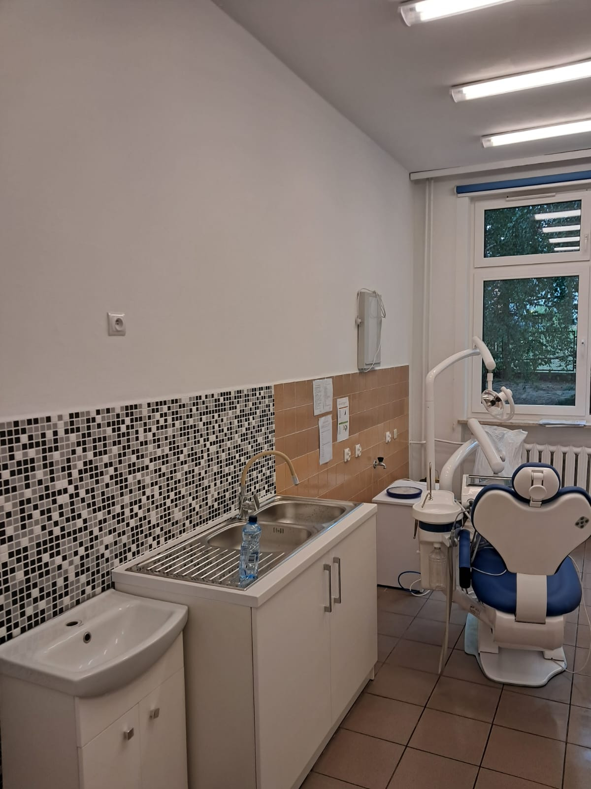 Umywalka z mozaiką w gabinecie dentystycznym, z niebieskim fotelem dentystycznym w tle. Widok na okno i zieleń na zewnątrz. Dominuje biel i jasne kolory.