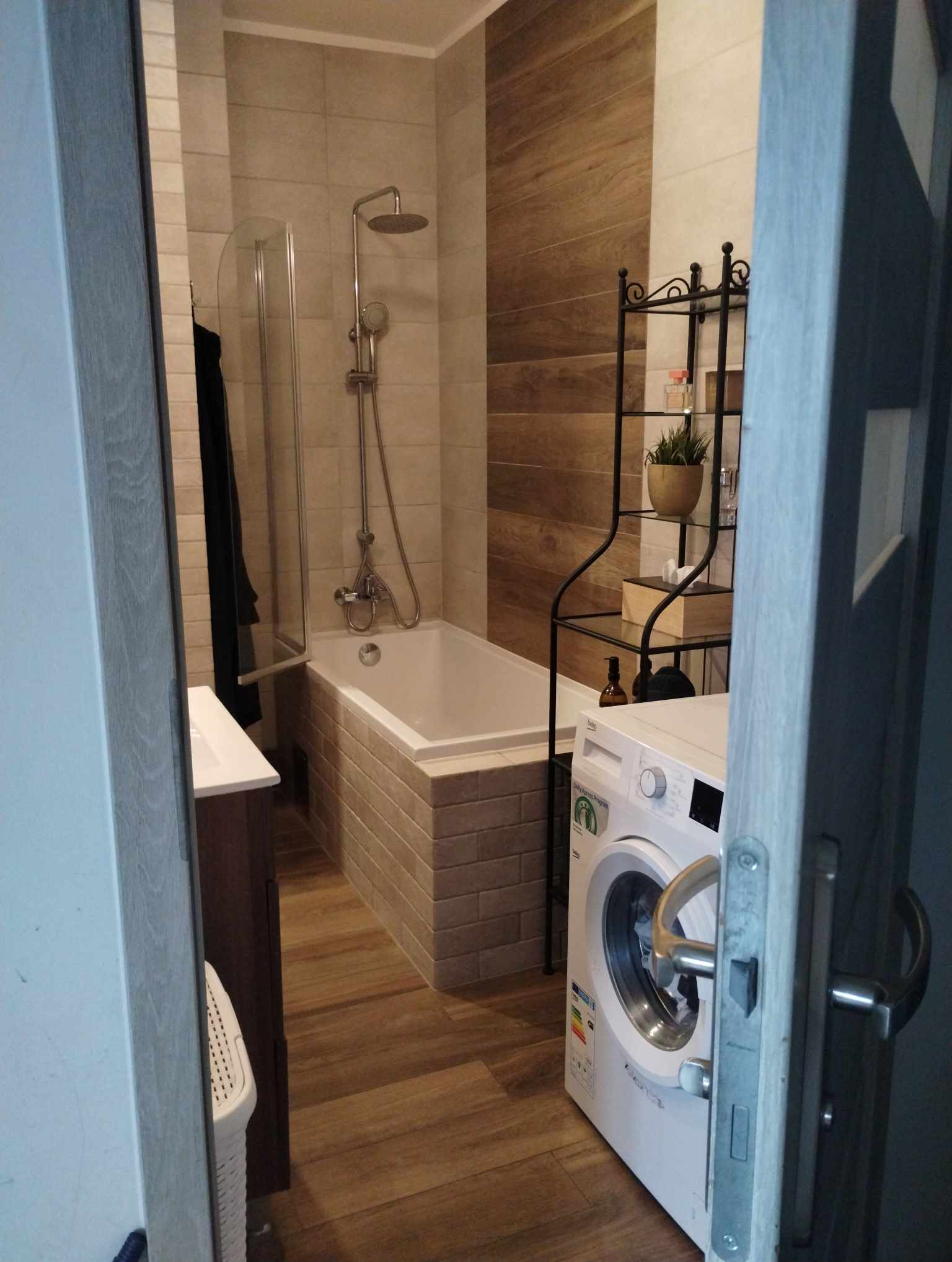 Wykończona łazienka z wanną obudowaną cegłą, kabiną prysznicową, pralką i drewnopodobnymi płytkami na podłodze i ścianie.