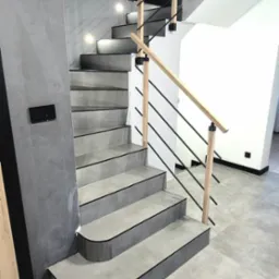 Nowoczesne schody z szarymi płytkami i drewnianą balustradą z czarnymi elementami, oświetlone punktowo, na tle szarej ściany i podłogi wyłożonej płytkami.
