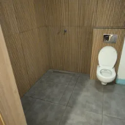 Nowoczesna łazienka z płytkami imitującymi drewno na ścianach i szarą podłogą. Widoczna toaleta z chromowanym przyciskiem spłukiwania i fragment ramy drzwi.