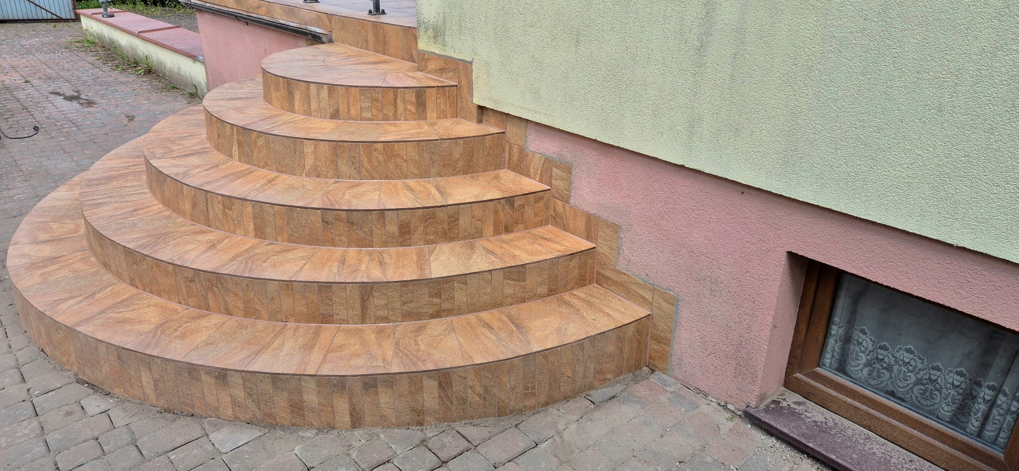 Wykończone klinkierem schody zewnętrzne, półokrągłe, z brązowymi płytkami, z widocznym oknem piwnicznym w tle. Detal wykończenia.