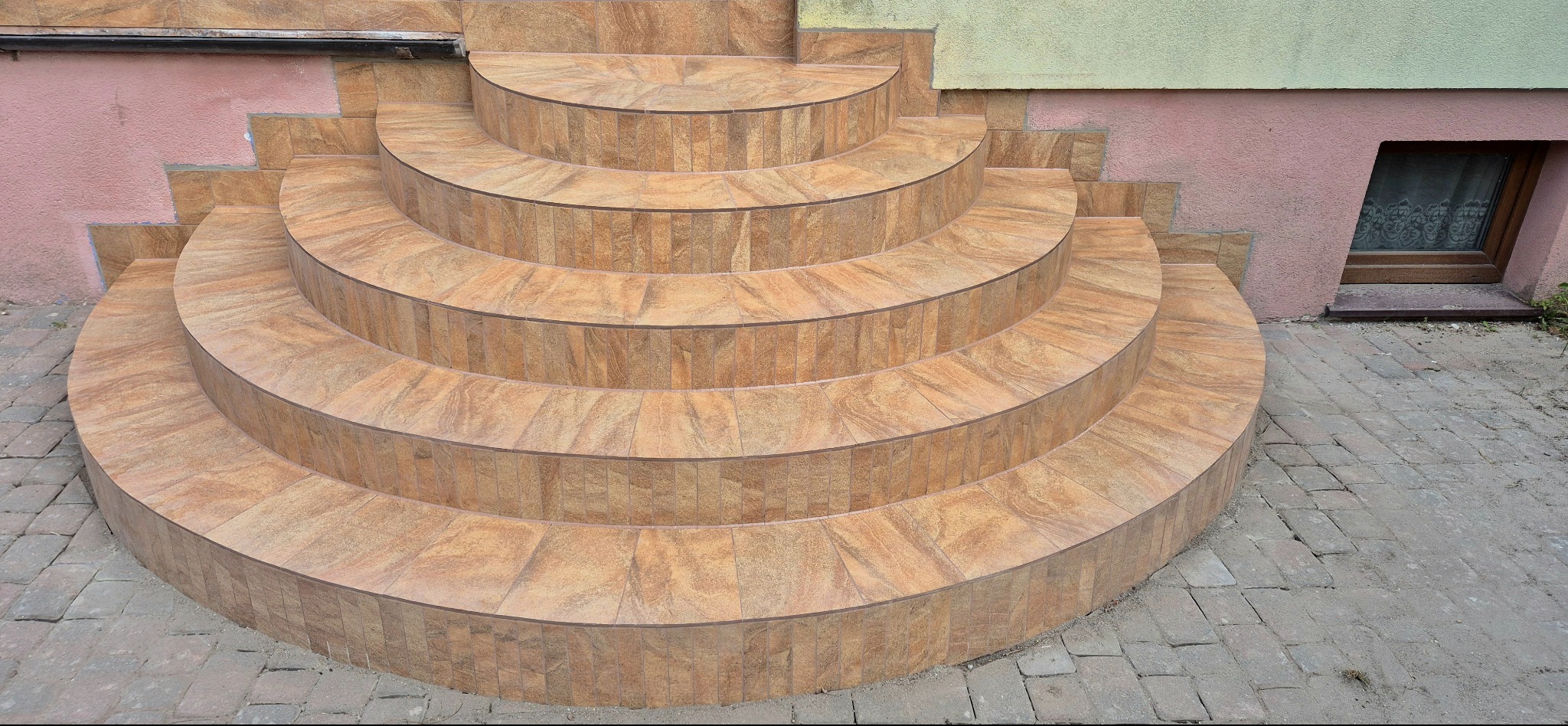 Półokrągłe schody wejściowe wyłożone beżową terakotą na zewnątrz budynku z oknem piwnicznym. Widoczne nierówne płytki chodnikowe i fragment elewacji w kolorze różowym i zielonym.