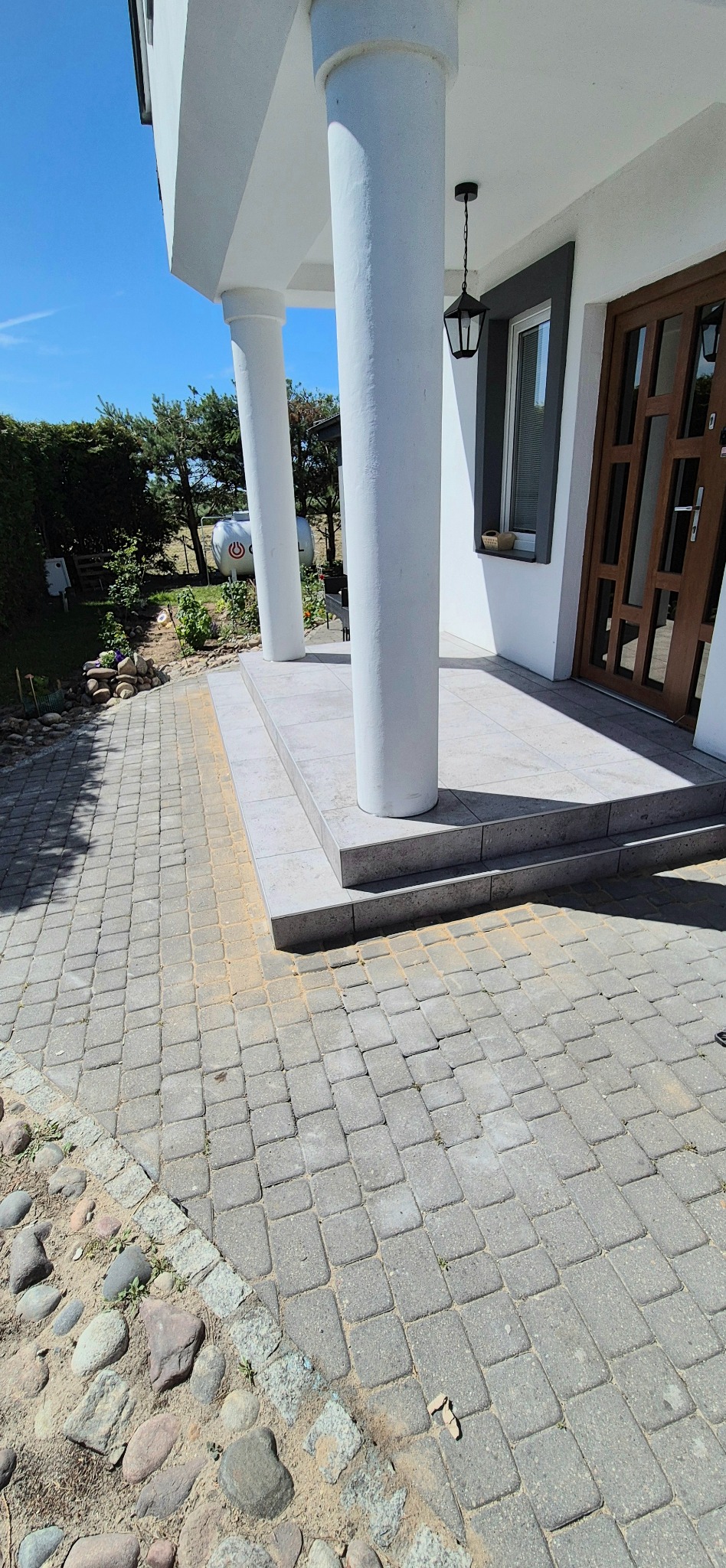 Wykończony taras z kolumnami i kostką brukową. Widoczny fragment domu z oknem i drzwiami. Jasne, słoneczne oświetlenie, błękitne niebo w tle.