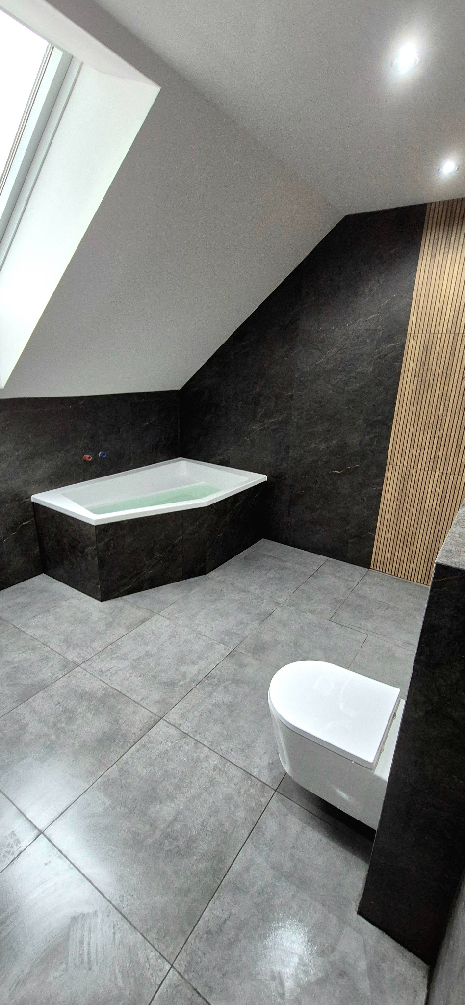 Nowoczesna łazienka na poddaszu z oknem dachowym, szarą płytką podłogową i ciemną okładziną ścienną imitującą kamień, uzupełniona o drewniane lamele.