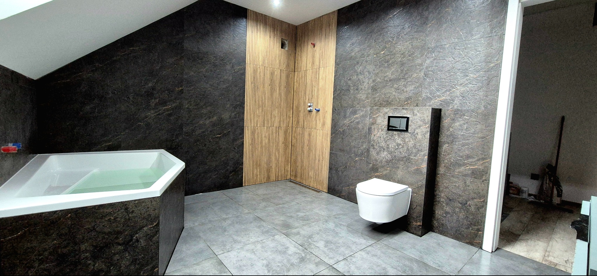 Nowoczesna łazienka z kabiną prysznicową wyłożoną drewnopodobnymi panelami, ciemnymi marmurkowymi płytkami i podwieszaną toaletą. Uwagę zwraca sześciokątna wanna.