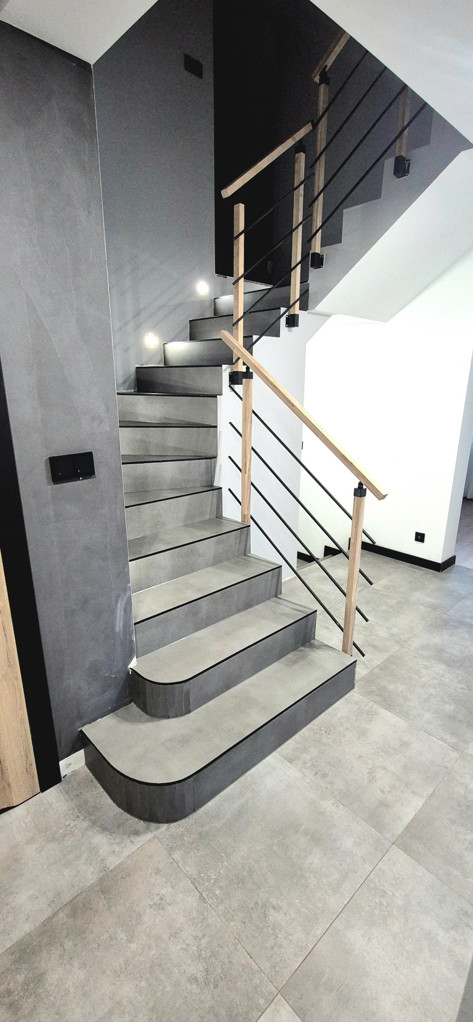 Nowoczesne schody z szarymi płytkami i drewnianą balustradą z czarnymi elementami, oświetlone punktowo, na tle szarej ściany i podłogi wyłożonej płytkami.