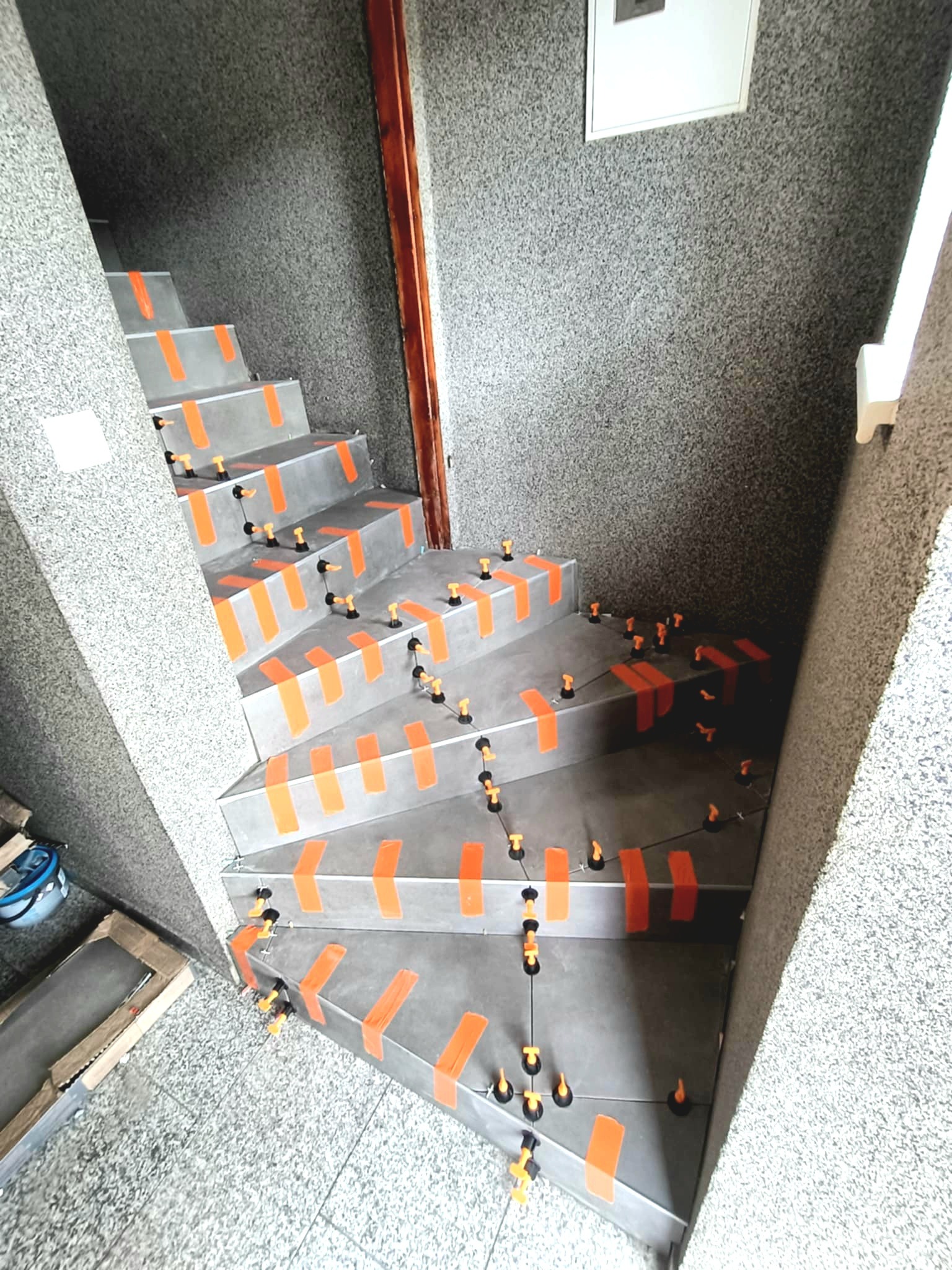 Schody w trakcie układania płytek, zabezpieczone pomarańczową taśmą i klipsami poziomującymi. Szara ściana z fakturą. Widoczne narzędzia glazurnicze.