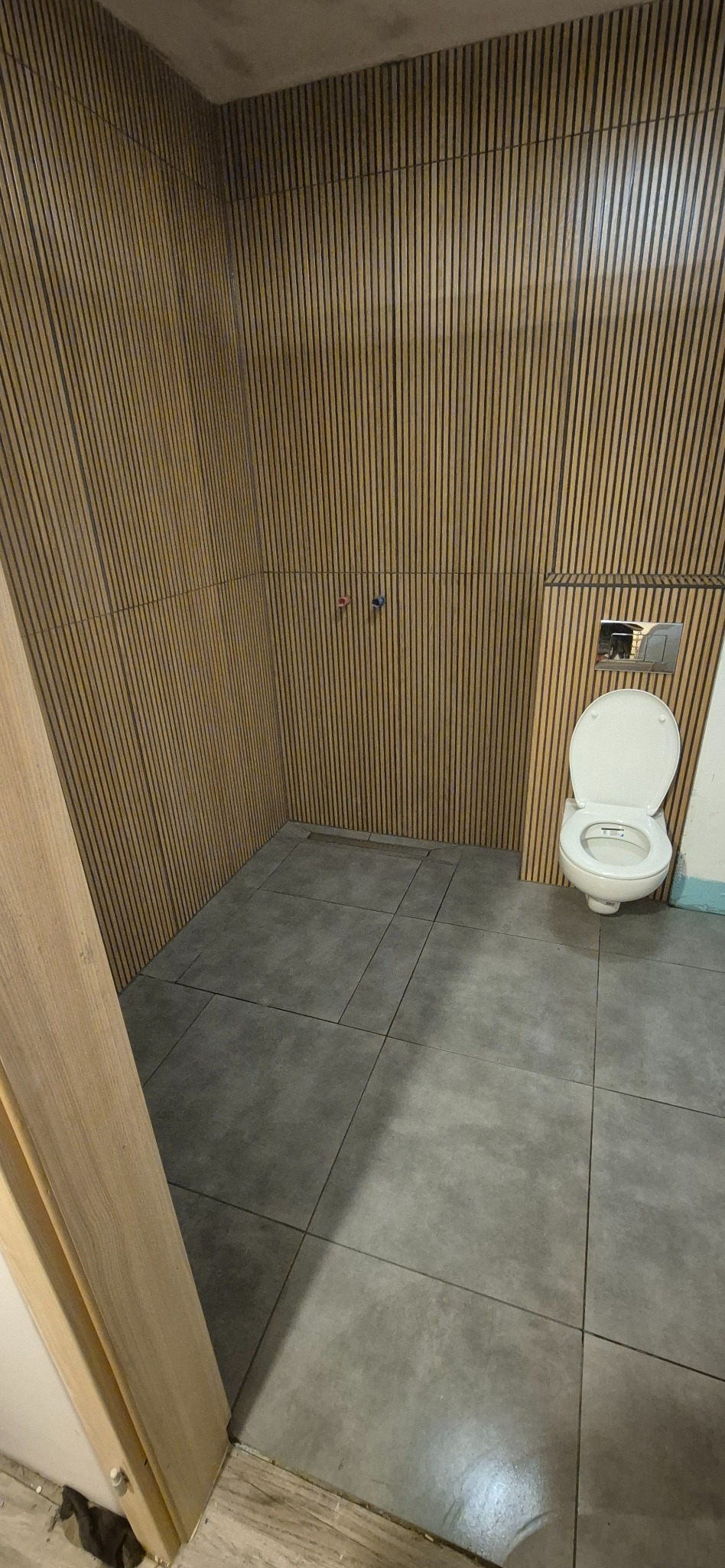 Nowoczesna łazienka z płytkami imitującymi drewno na ścianach i szarą podłogą. Widoczna toaleta z chromowanym przyciskiem spłukiwania i fragment ramy drzwi.