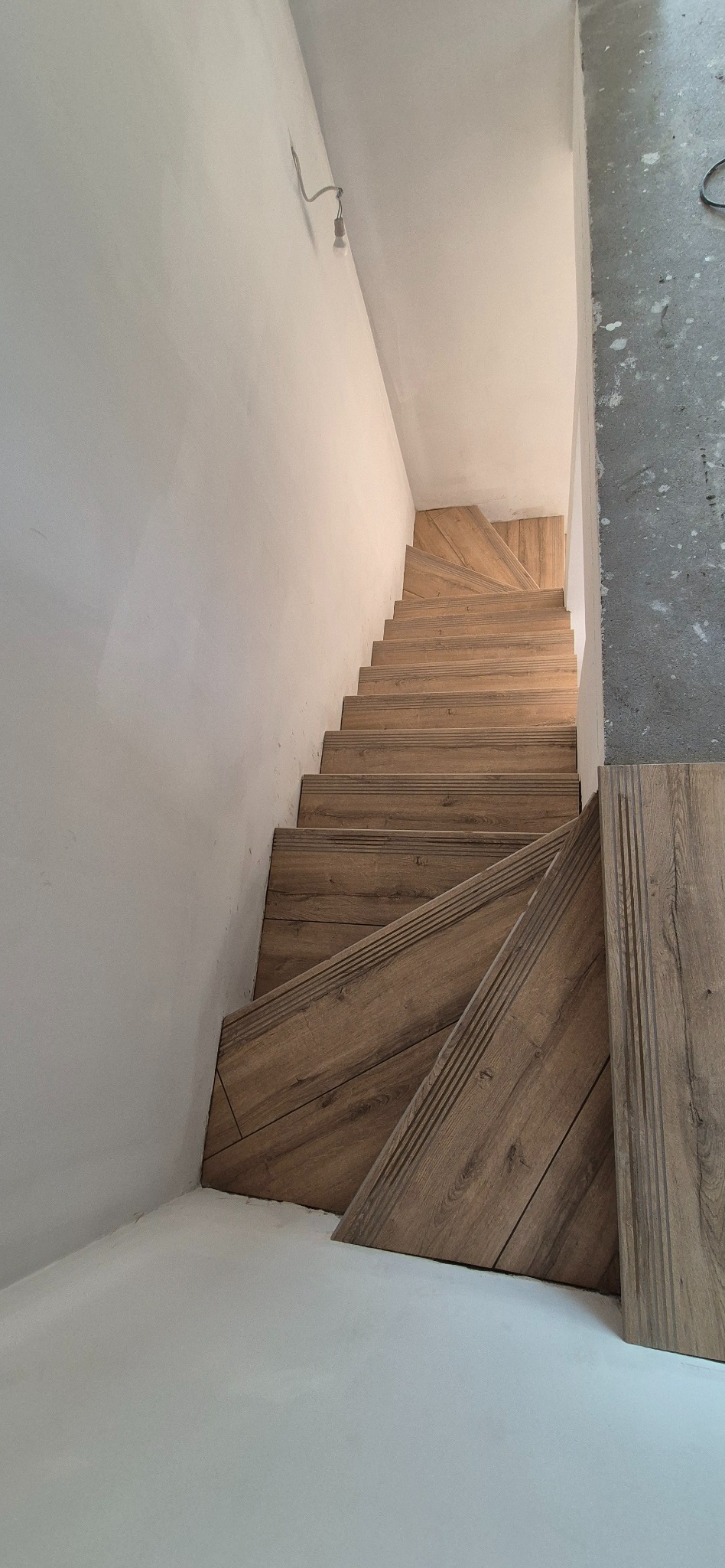 Wykończone schody z drewnopodobnych paneli, widok z góry. Fragment ściany z surowego betonu. Oświetlenie sufitowe w tle.
