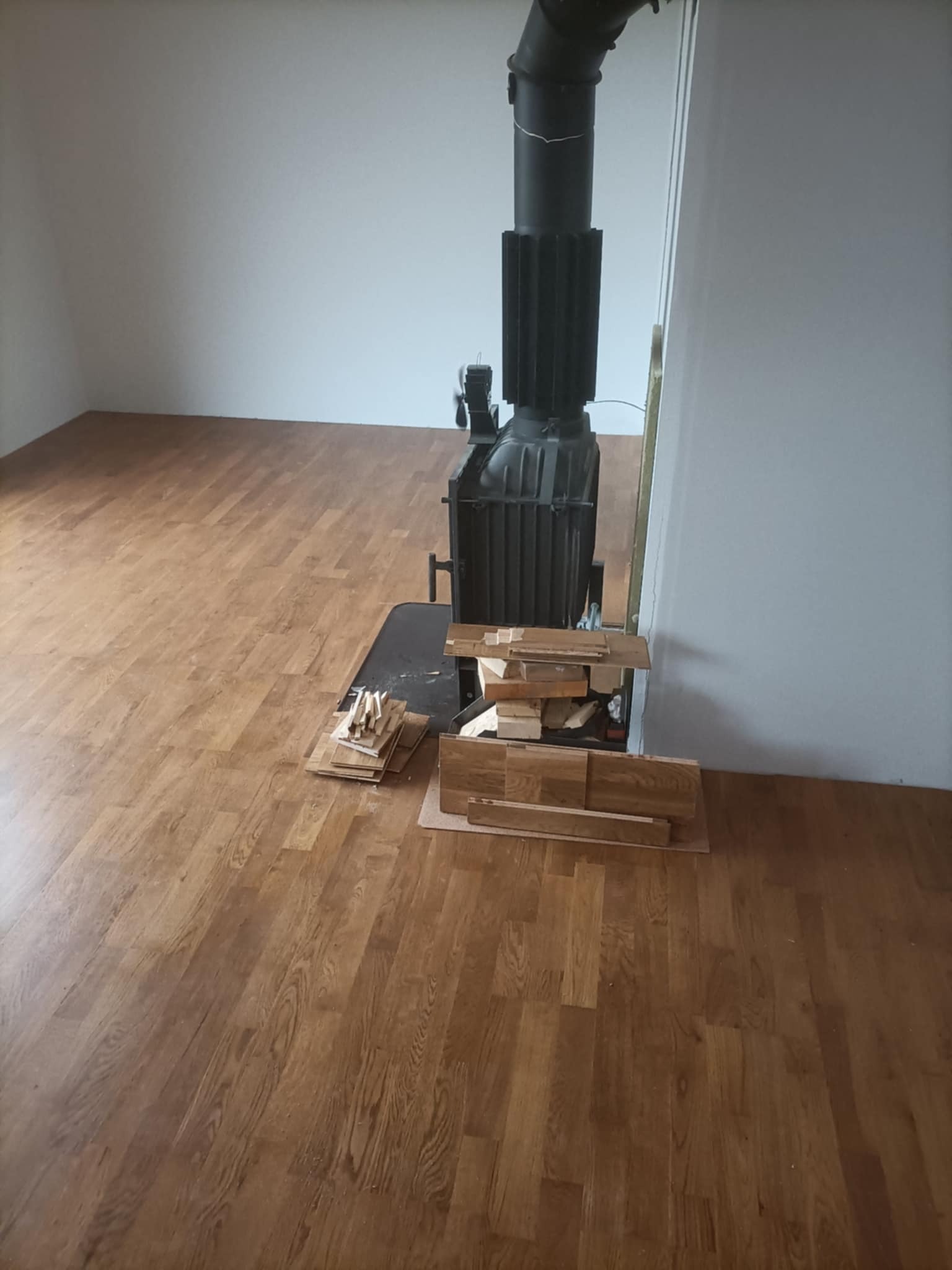 Ułożony parkiet w pokoju z kominkiem; deski podłogowe leżą obok kominka, przygotowane do dalszego montażu. Widoczne jasne ściany i brązowa podłoga.