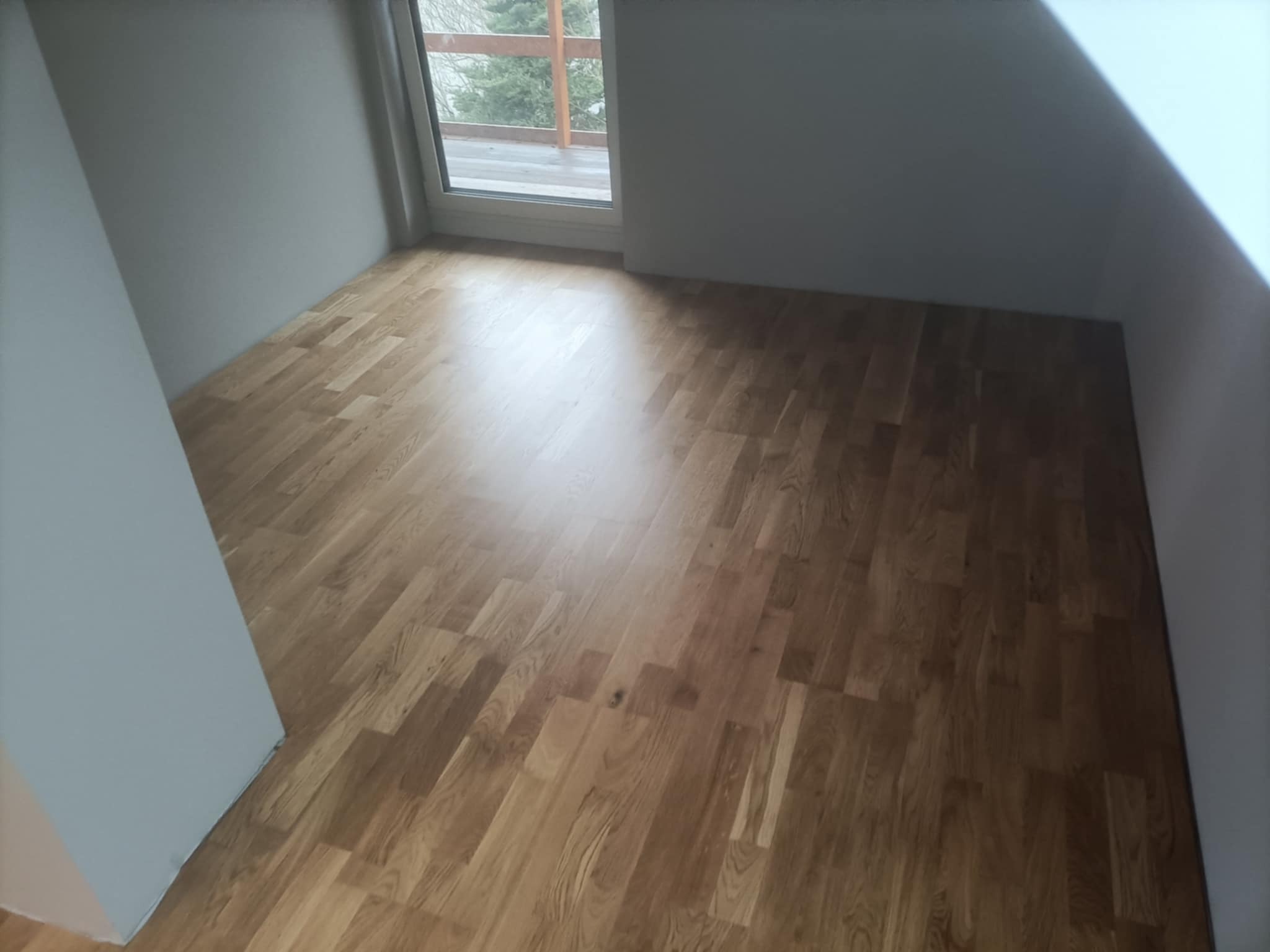 Jasny parkiet ułożony w pokoju na poddaszu, widoczne okno balkonowe i skosy. Świeżo wykończona podłoga z naturalnego drewna, oddająca ciepło wnętrza.