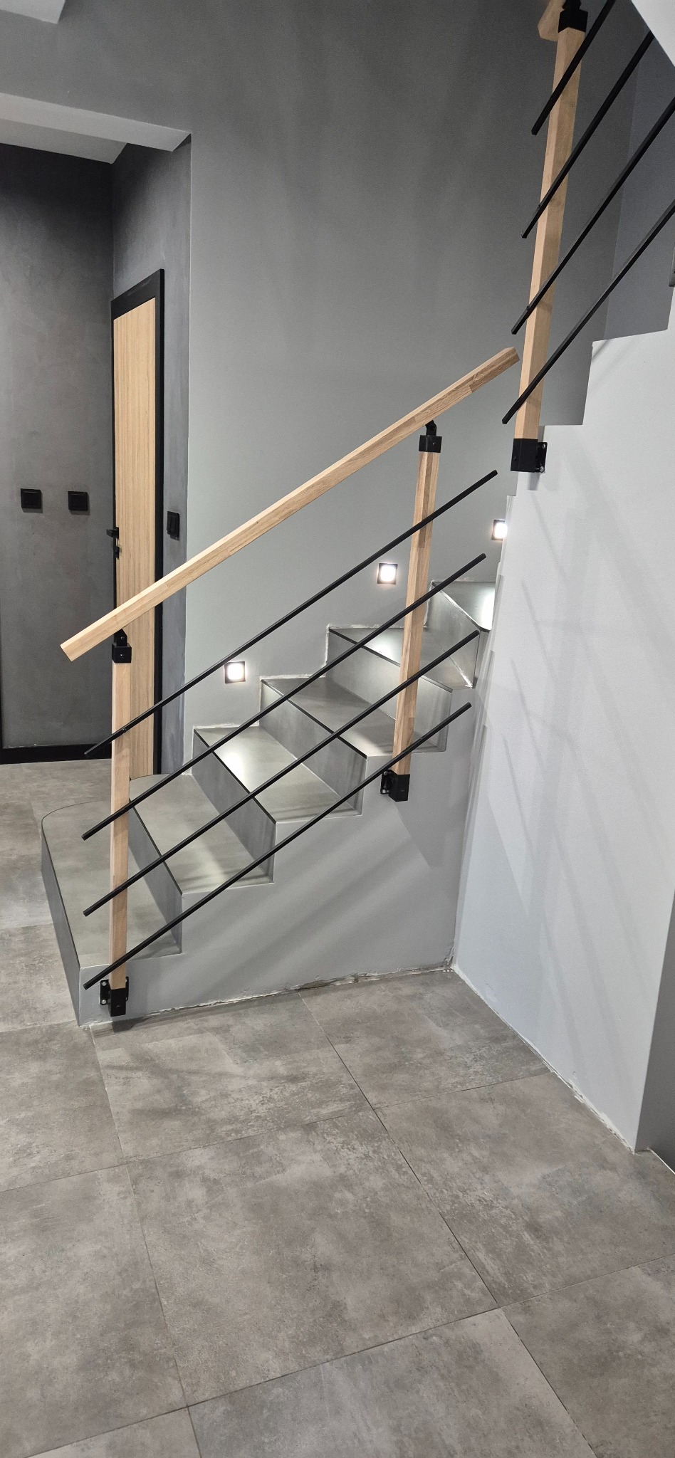 Nowoczesne schody z drewnianą balustradą i czarnymi metalowymi elementami, oświetlone punktowymi lampami, na tle szarej ściany i podłogi z płytek.