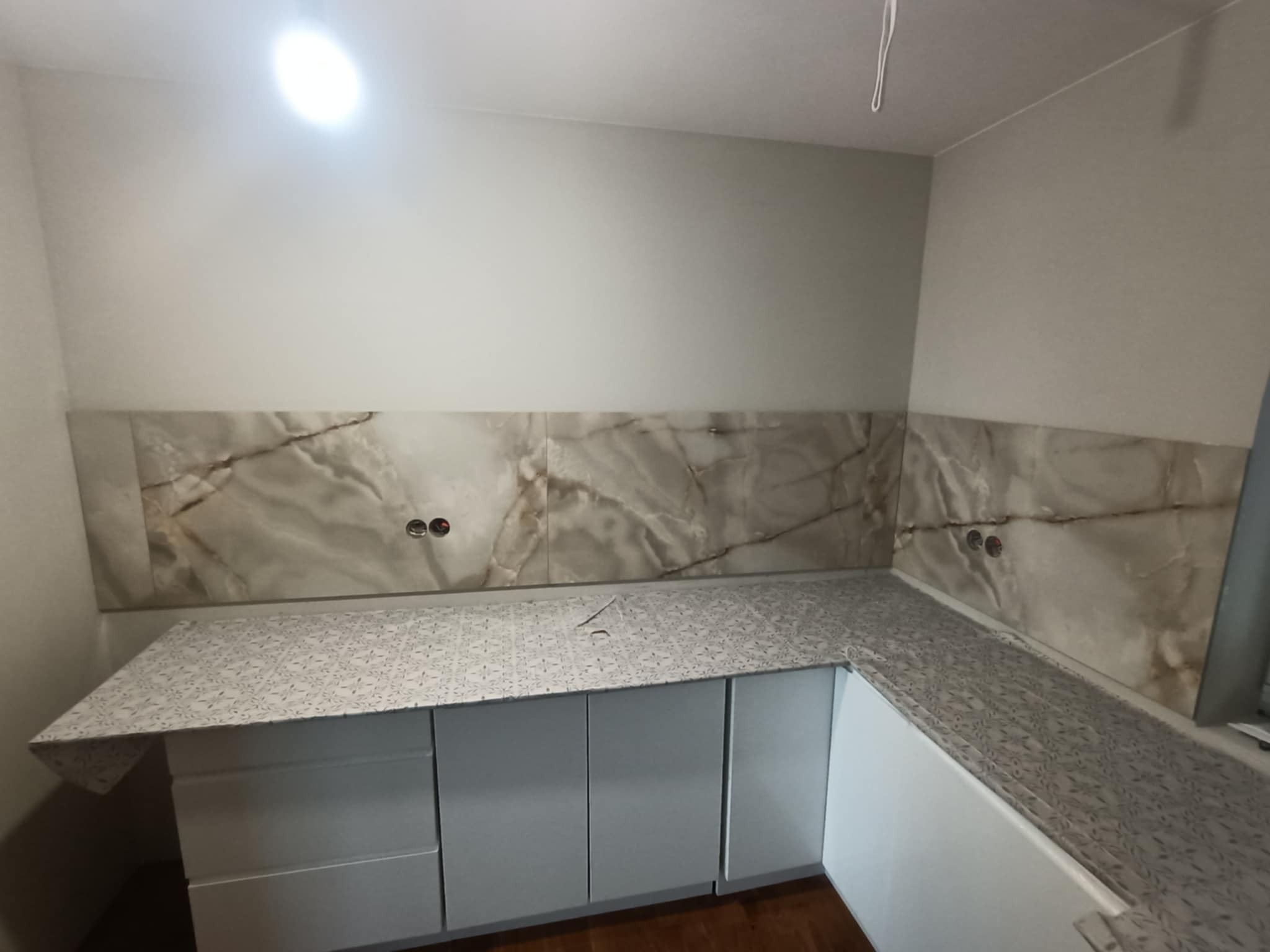 Wykończona kuchnia z marmurowym panelem nad blatem i jasnoszarymi szafkami. Blat z subtelnym wzorem. Widoczne gniazdka elektryczne.