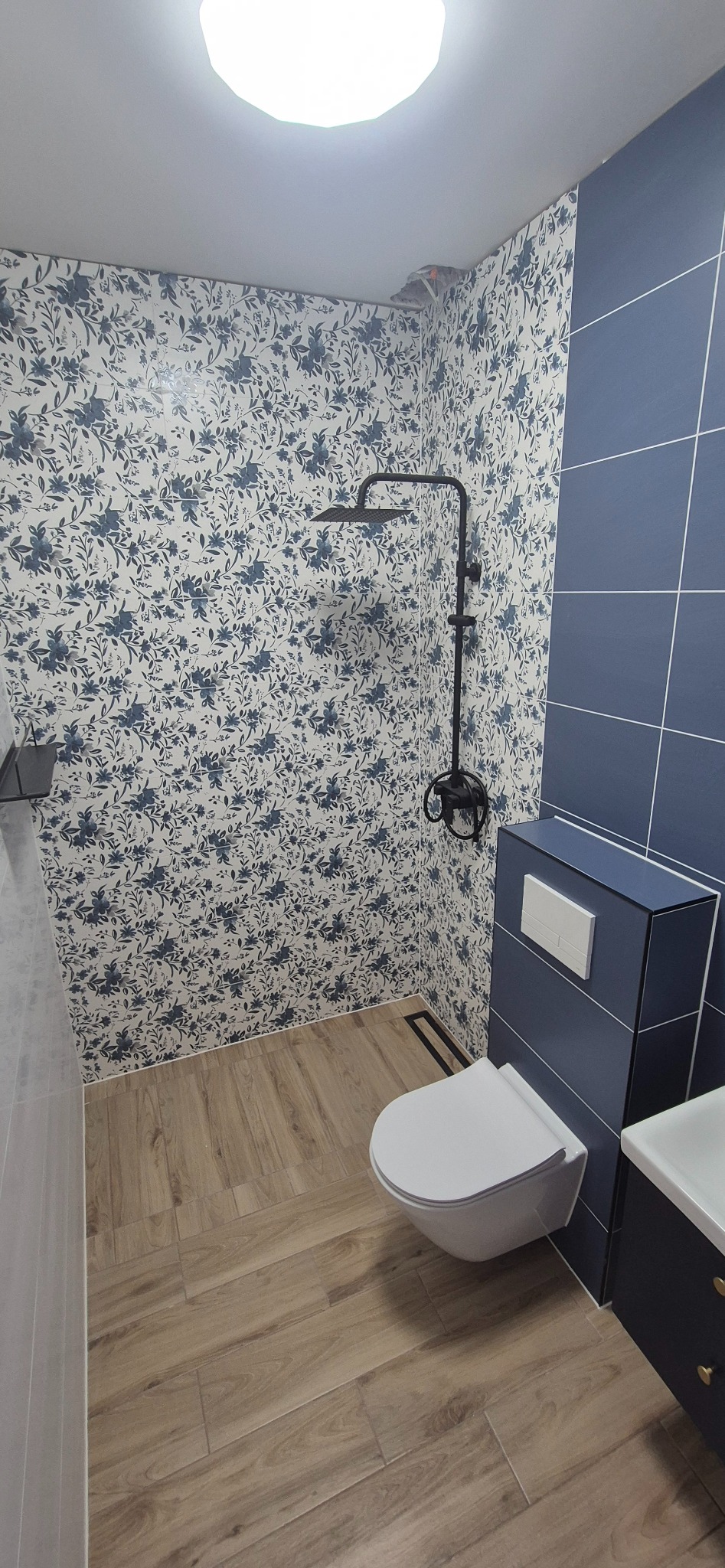 Wykończona łazienka z toaletą podwieszaną, kabiną prysznicową z czarną armaturą i drewnopodobnymi płytkami podłogowymi. Ściany wyłożone płytkami w kwiatowy wzór i niebieskimi płytkami.