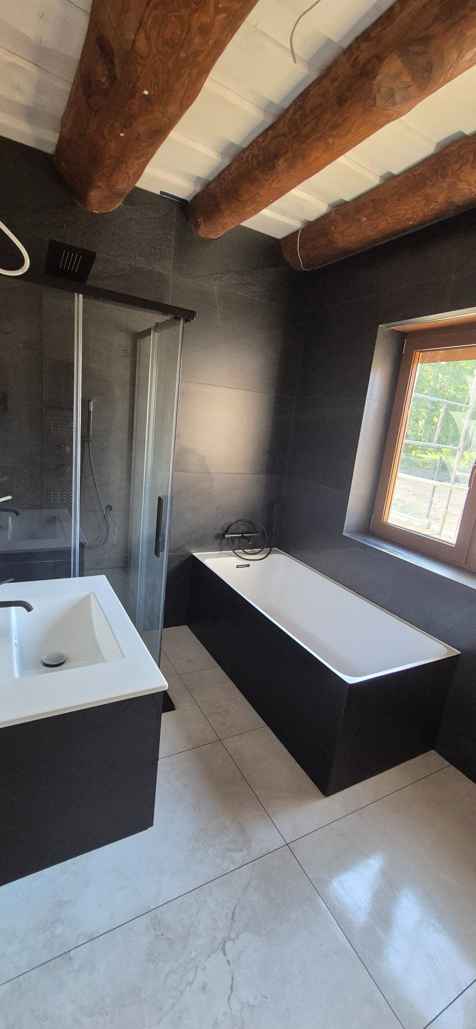 Nowoczesna łazienka z czarnymi płytkami, kabiną prysznicową, wanną i drewnianymi belkami stropowymi. Minimalistyczny design z naturalnym oświetleniem.