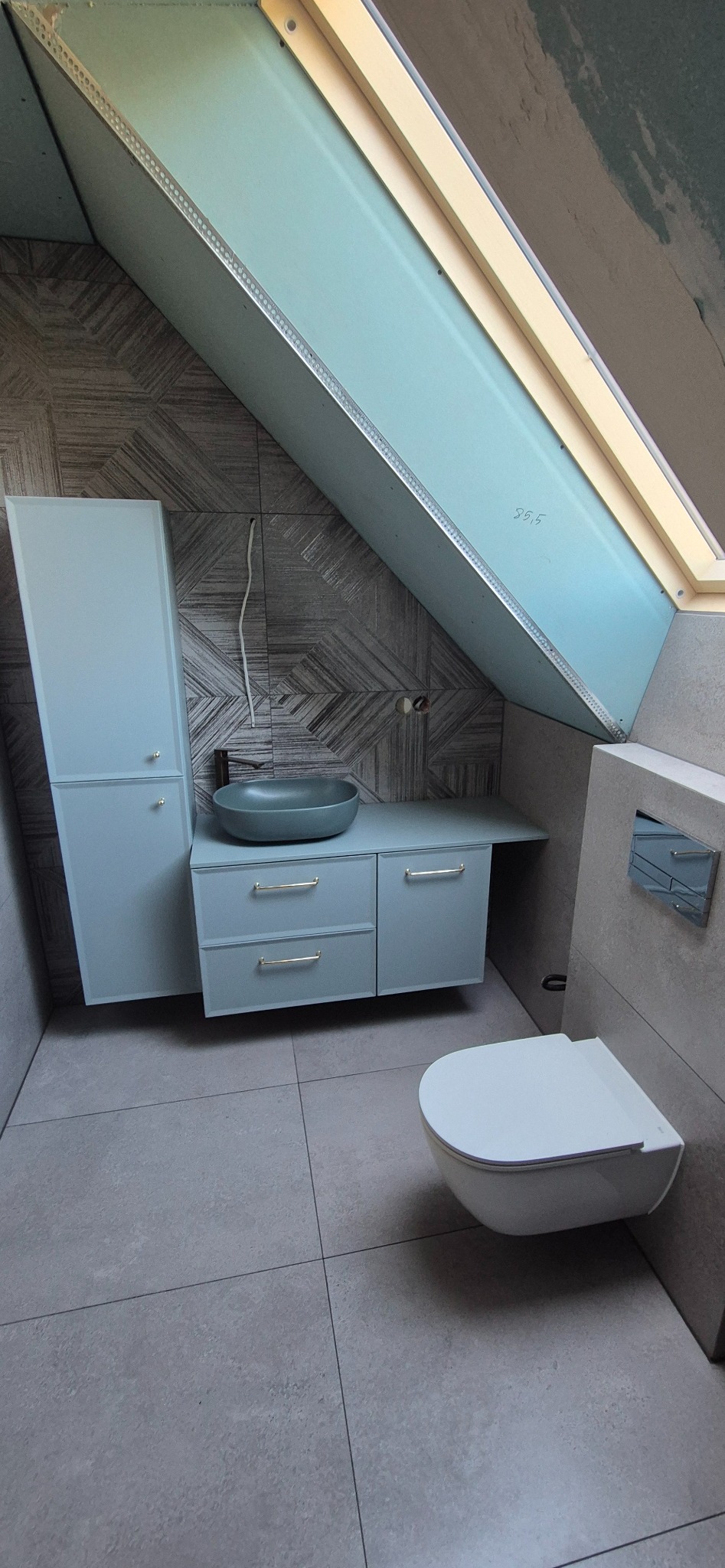 Łazienka na poddaszu z umywalką, toaletą i szafką w odcieniach szarości. Ukośne ściany wykończone płytkami o geometrycznym wzorze. Widoczne okno dachowe.