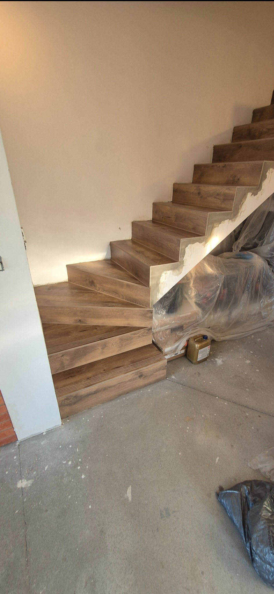 Montaż drewnopodobnych płytek na schodach w trakcie prac wykończeniowych. Widoczne zabezpieczenia i materiały budowlane. Ujęcie z dołu schodów.