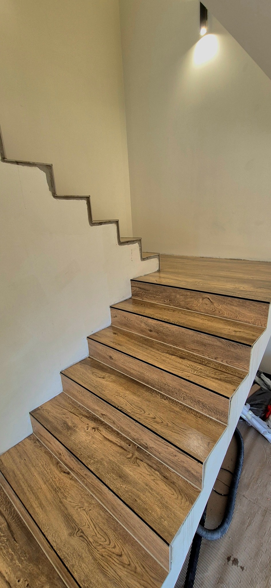 Wykończone schody z płytek imitujących drewno, z czarnymi fugami, na tle jasnej ściany z widocznymi śladami po schodach. Widok z dołu, oświetlenie górne.