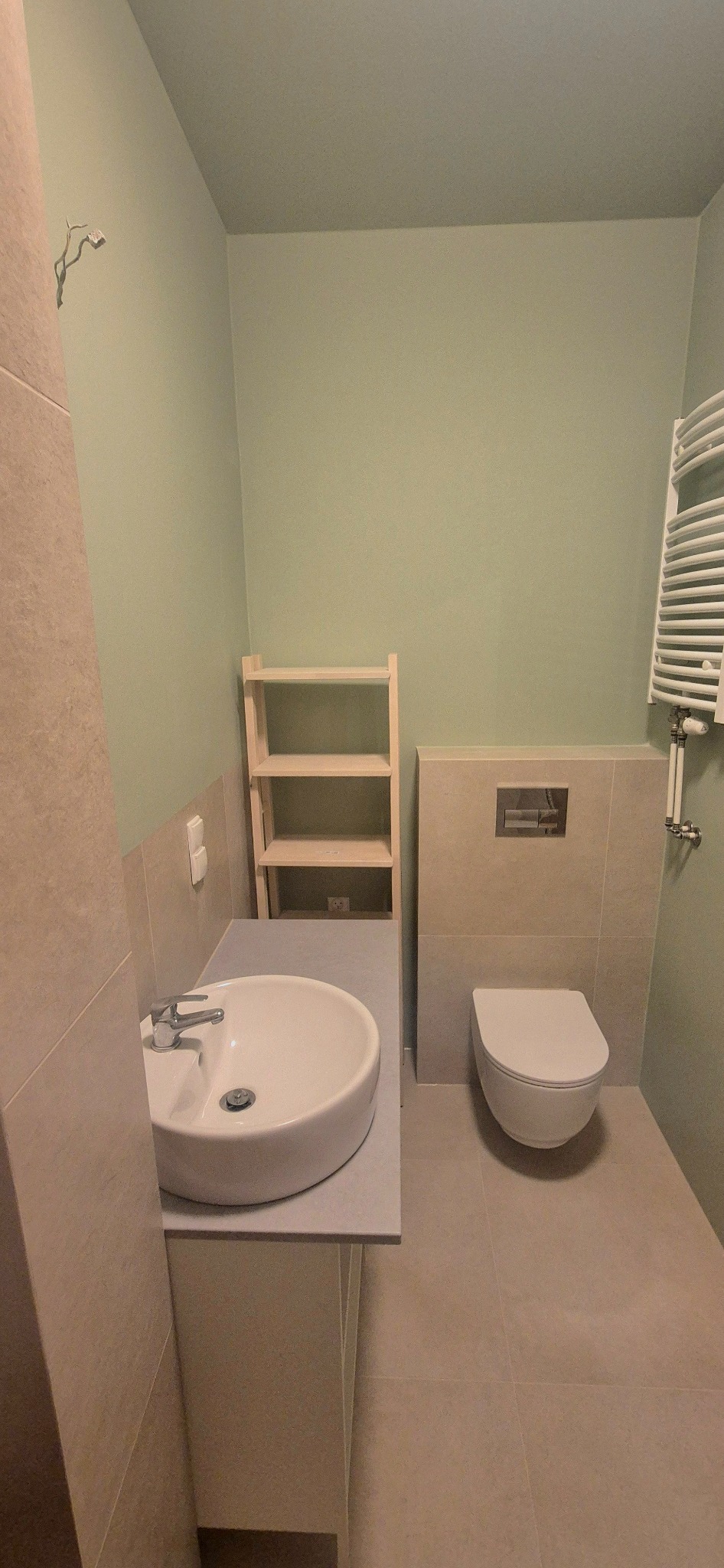 Jasna łazienka z umywalką, toaletą i grzejnikiem drabinkowym. Ściany w odcieniach beżu i zieleni. Widoczna drewniana półka. Minimalistyczny styl.