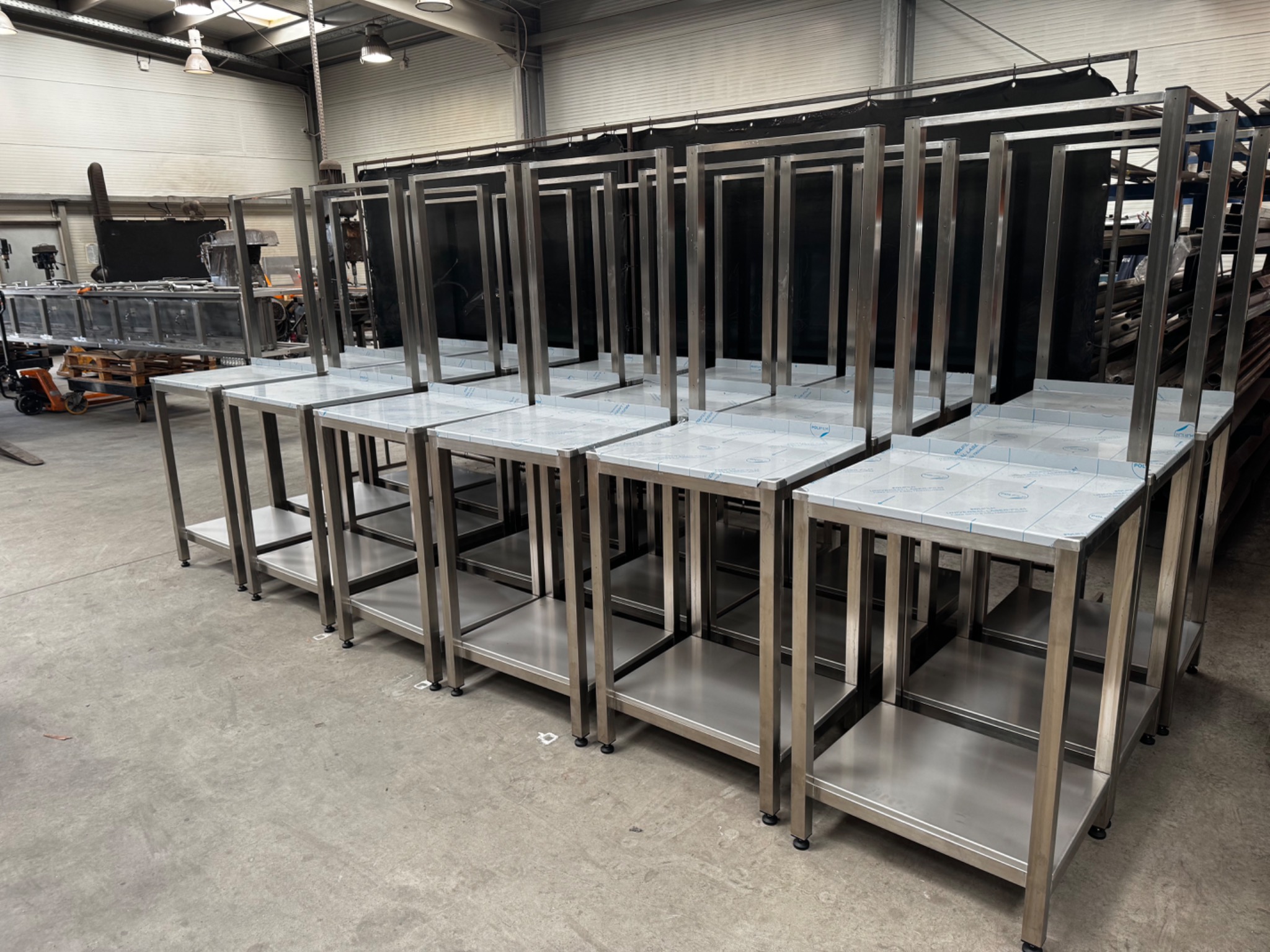 Rząd identycznych, spawanych stołów ze stali nierdzewnej z blatem pokrytym folią ochronną, ustawionych w hali produkcyjnej.