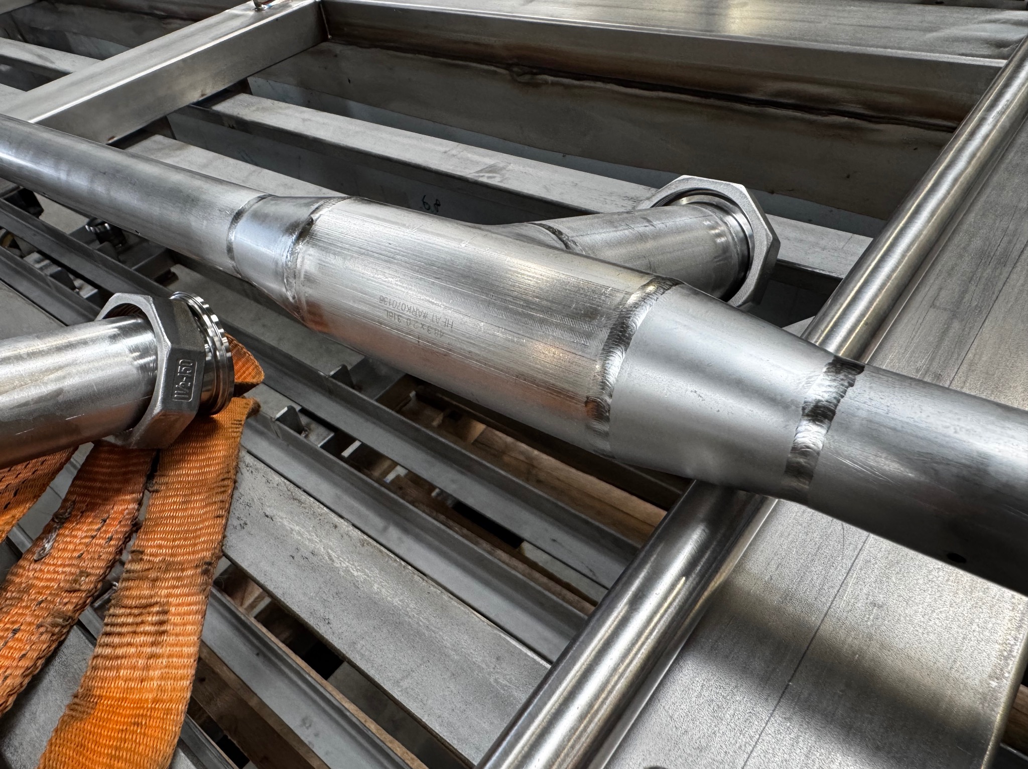 Szczegółowe ujęcie spawanych, nierdzewnych rur i konstrukcji stalowej z pomarańczowym pasem transportowym. Usługi spawacza w Szczecinie.