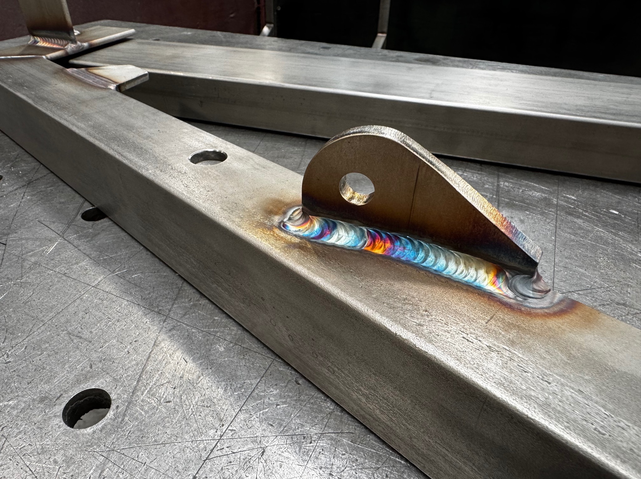 Metalowy element konstrukcyjny z precyzyjnym spoiwem spawalniczym, widoczne tęczowe przebarwienia na powierzchni spoiny. Detal konstrukcji stalowej z bliska.