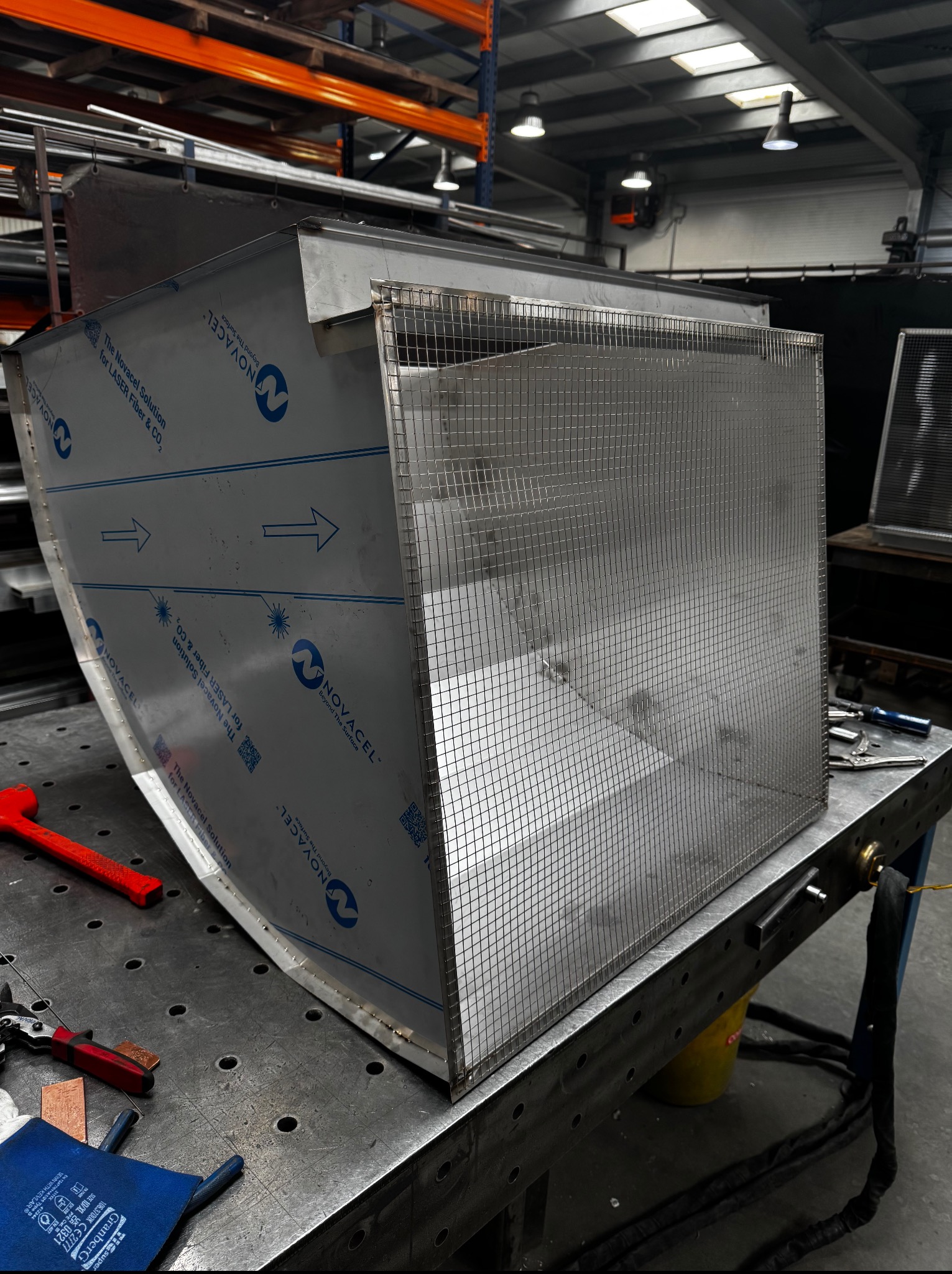 Konstrukcja z blachy nierdzewnej i siatki na stole warsztatowym, widoczne narzędzia i elementy montażowe, hala produkcyjna w tle. Detal spawalniczy w trakcie realizacji.