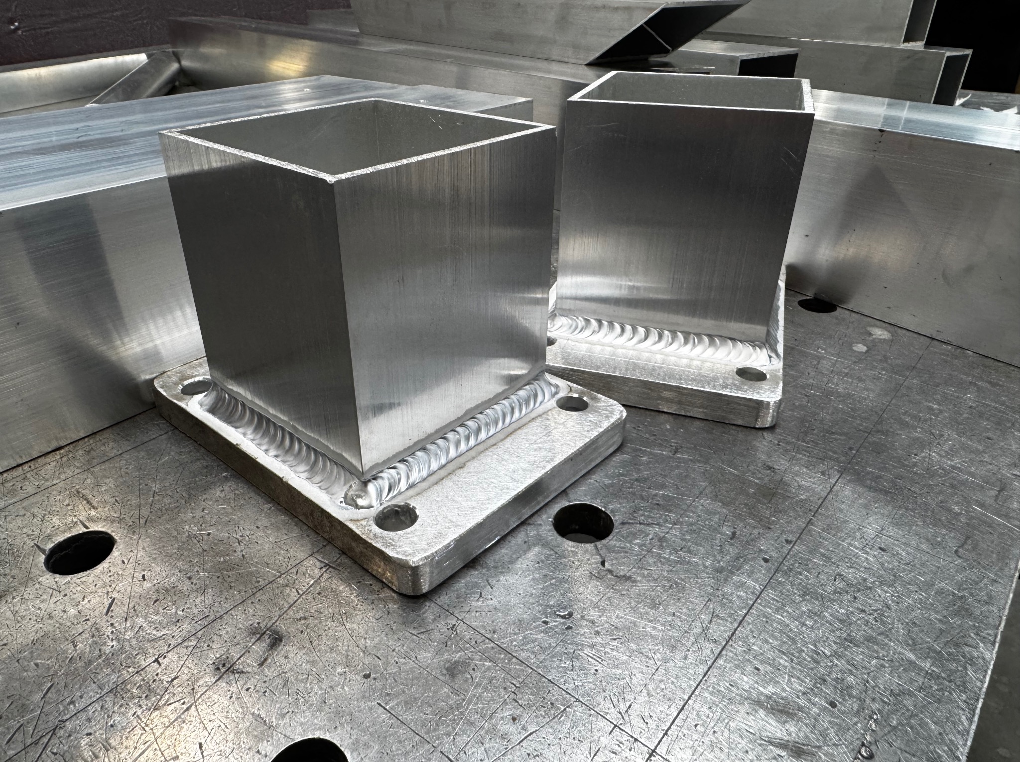 Dwa spawane, aluminiowe profile kwadratowe na metalowym stole spawalniczym. Widoczne ślady spawania i obróbki metalu. Surowy, industrialny wygląd.