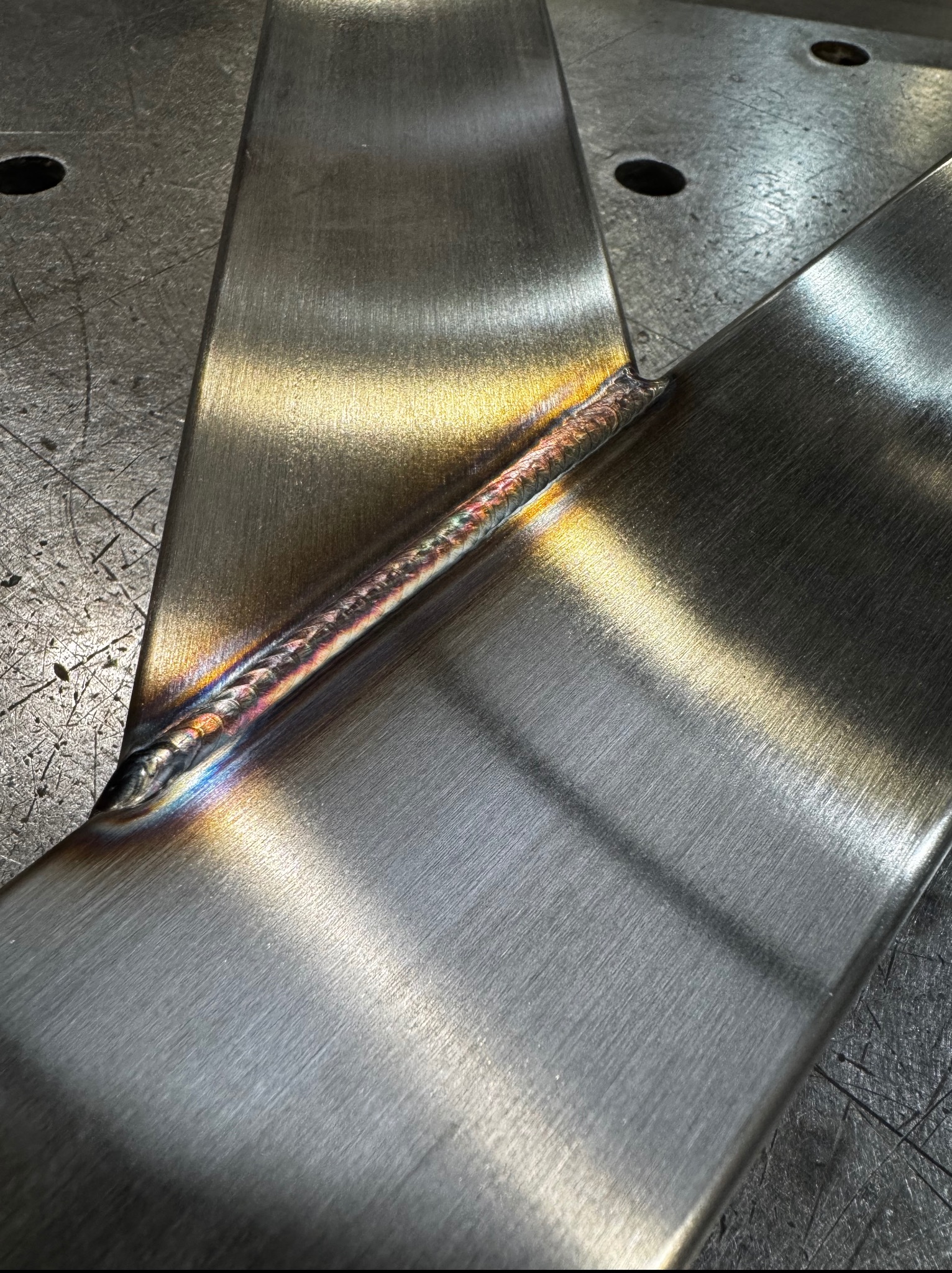 Detal spawu łączącego dwie metalowe płyty pod kątem. Widoczne tęczowe przebarwienia na spoinie, świadczące o wysokiej temperaturze i jakości wykonania.