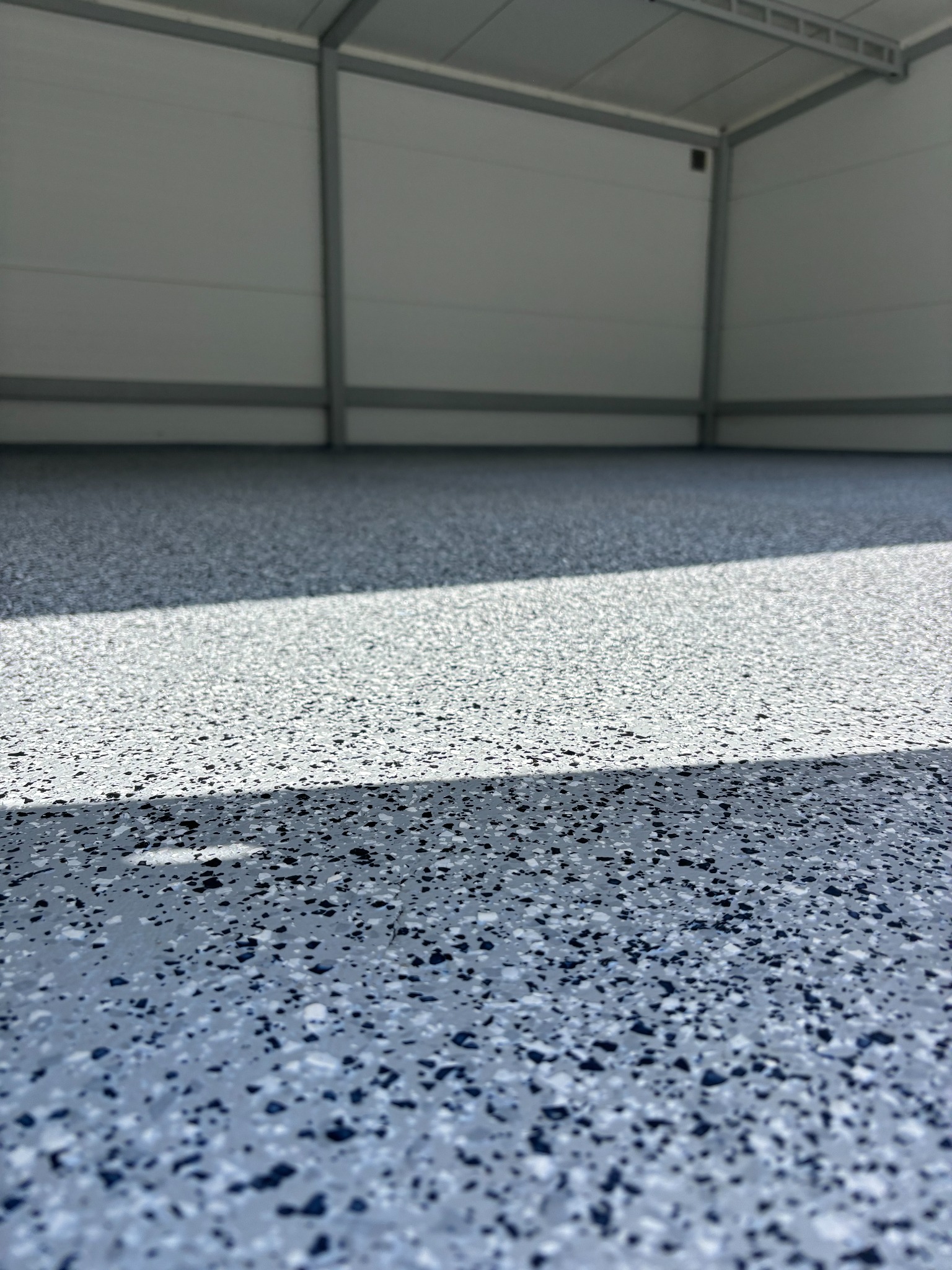 Szara posadzka z żywicy epoksydowej z czarnymi i białymi płatkami w hali magazynowej, oświetlona naturalnym światłem. Gładka powierzchnia z delikatnym połyskiem.