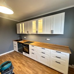 Oleksandr Urutskyi - Biała kuchnia z drewnianym blatem i czarnymi uchwytami, oświetlona lampą sufitową. Widoczny odkurzacz na podłodze. Szare ściany.