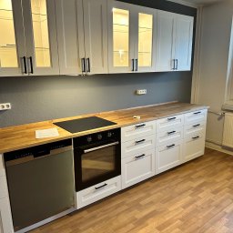 Oleksandr Urutskyi - Biała kuchnia z drewnianym blatem i czarnymi uchwytami, oświetlone szafki wiszące z przeszkleniami, płyta indukcyjna i piekarnik w zabudowie, widok na okno.