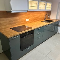 Oleksandr Urutskyi - Nowoczesna kuchnia z szarymi szafkami, drewnianym blatem i panelem ściennym. Widoczna płyta indukcyjna, piekarnik i czarny zlew. Górne szafki z podświetleniem.